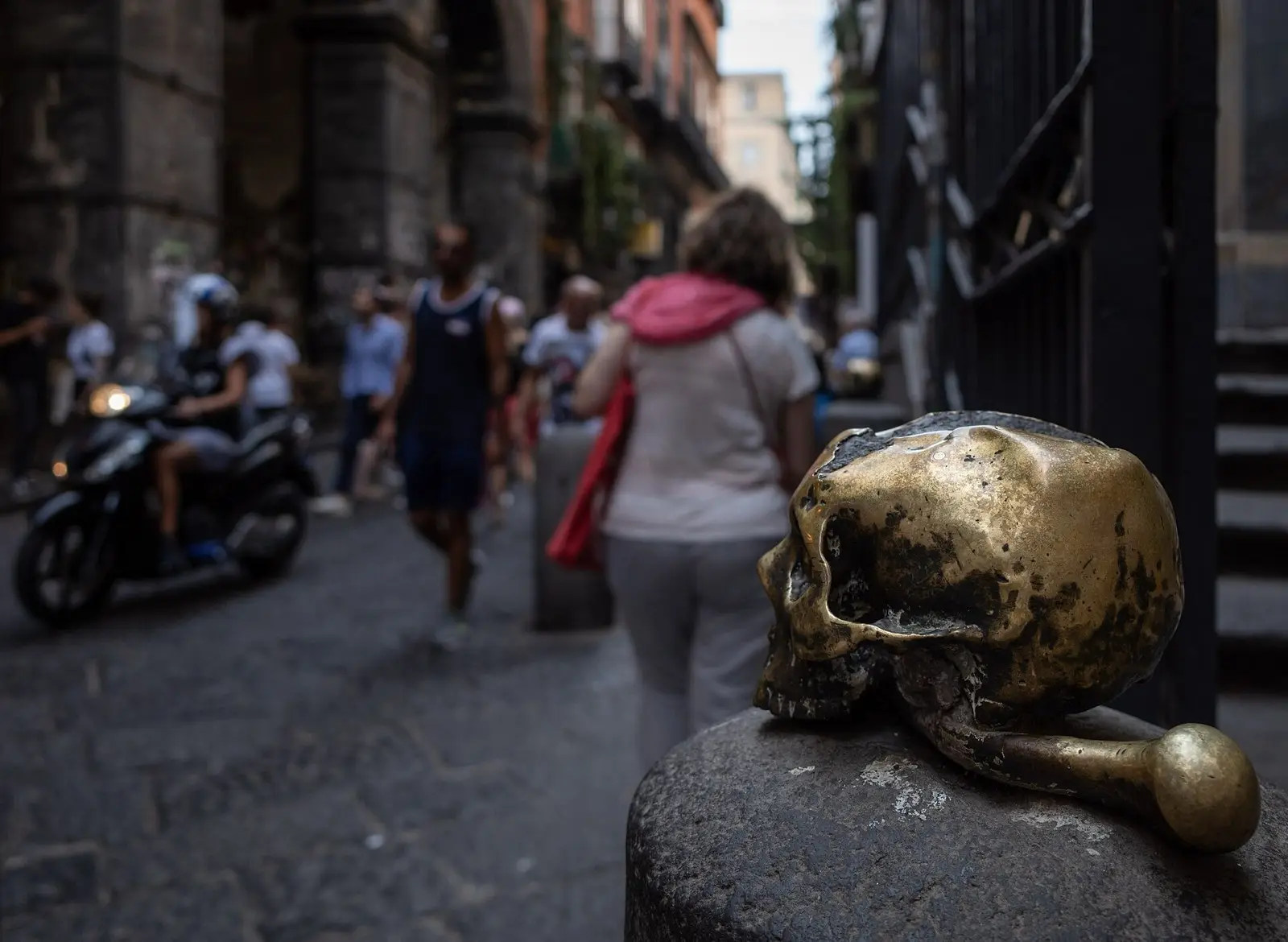 A polished bronze skull on a post outside the church of Purgatorio ad Arco, with people walking along Via dei Tribunali
