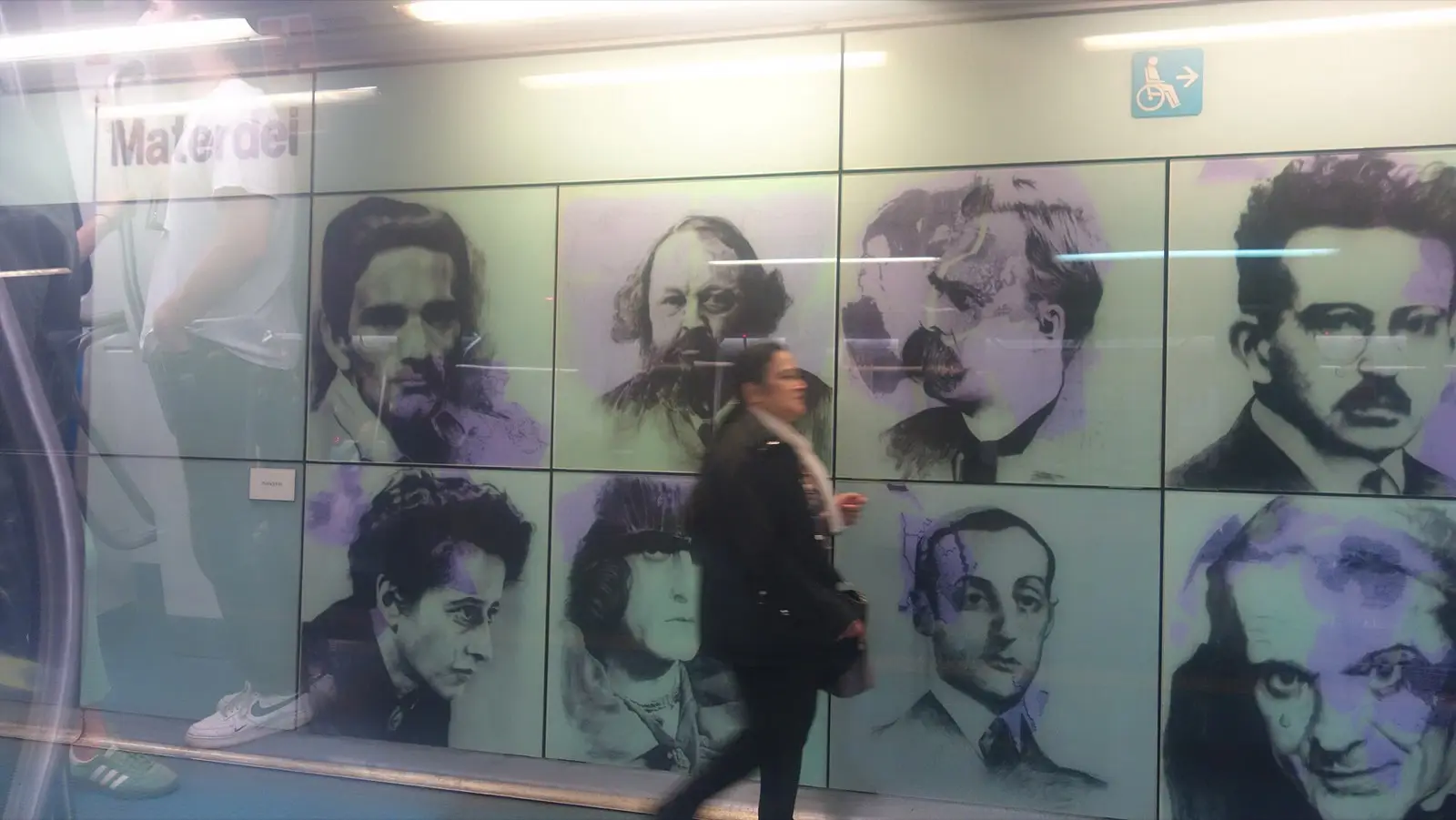 A wall of large printed portraits at Materdei station with a woman walking past
